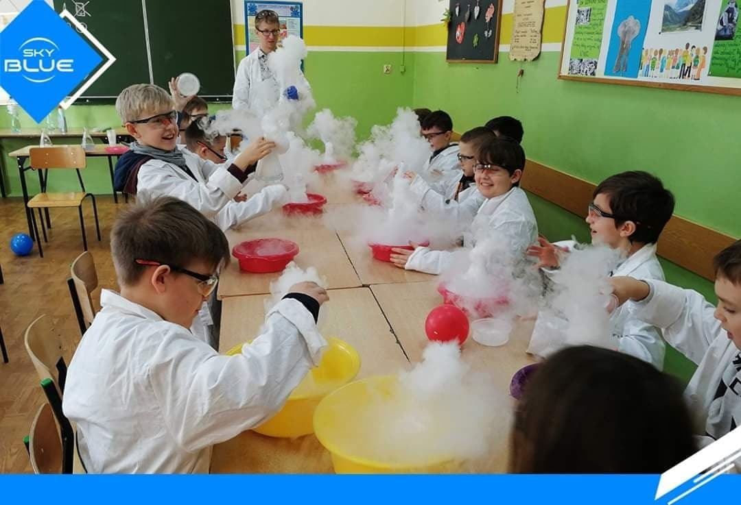 Grupa dzieci w białych fartuchach i okularach ochronnych przeprowadza eksperyment z suchym lodem w klasie, tworząc obłoki pary nad kolorowymi miskami.