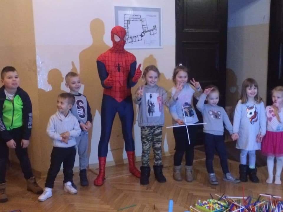 Grupa dzieci pozuje do zdjęcia z osobą przebraną za Spider-Mana na tle ściany z planem budynku, na podłodze leżą kolorowe patyczki do budowania.