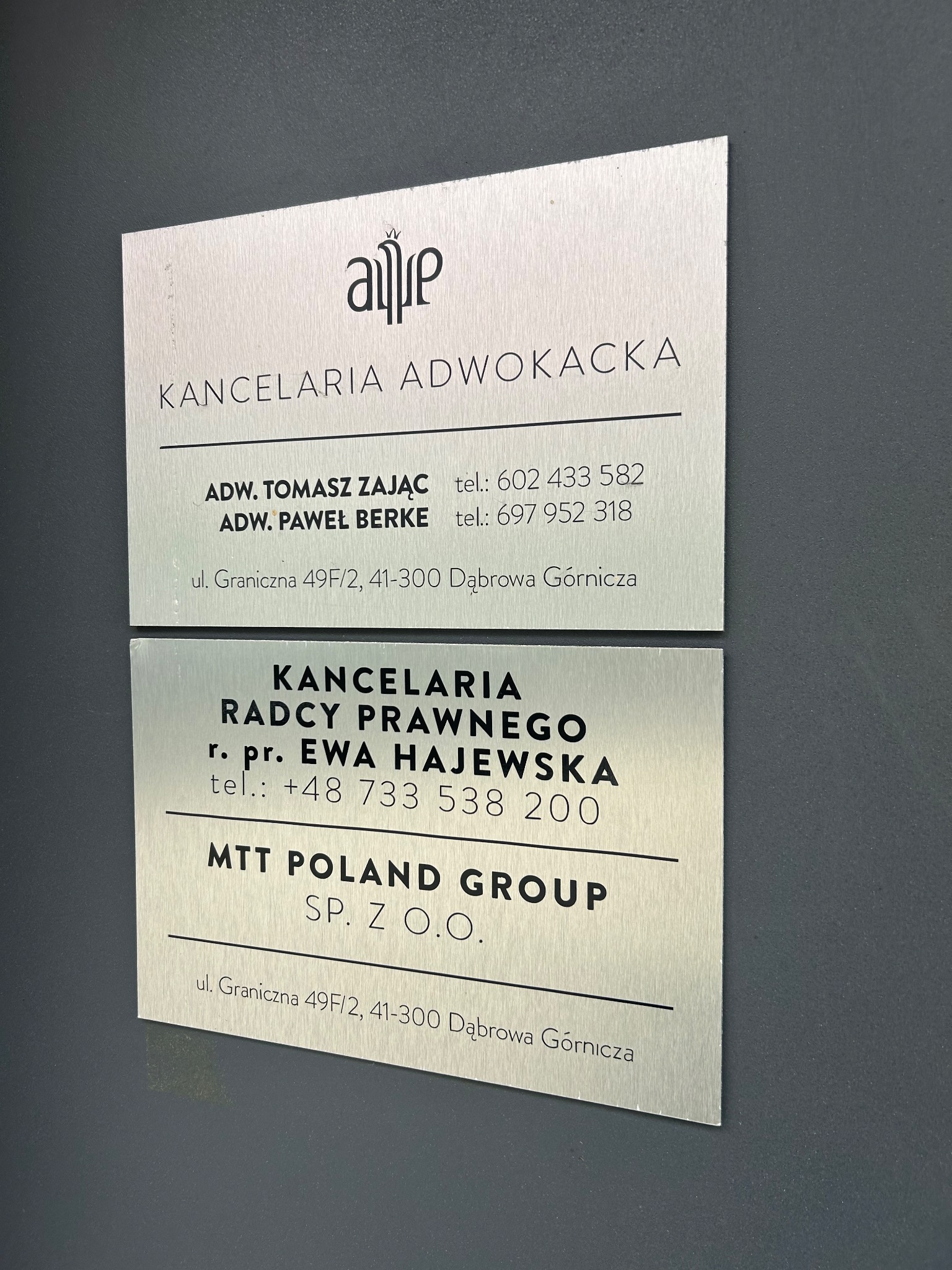 Dwie metalowe tabliczki informacyjne na ścianie, prezentujące Kancelarię Adwokacką Tomasza Zająca i Pawła Berke oraz Kancelarię Radcy Prawnego Ewy Hajewskiej, obie zlokalizowane w Dąbrowie Górniczej.