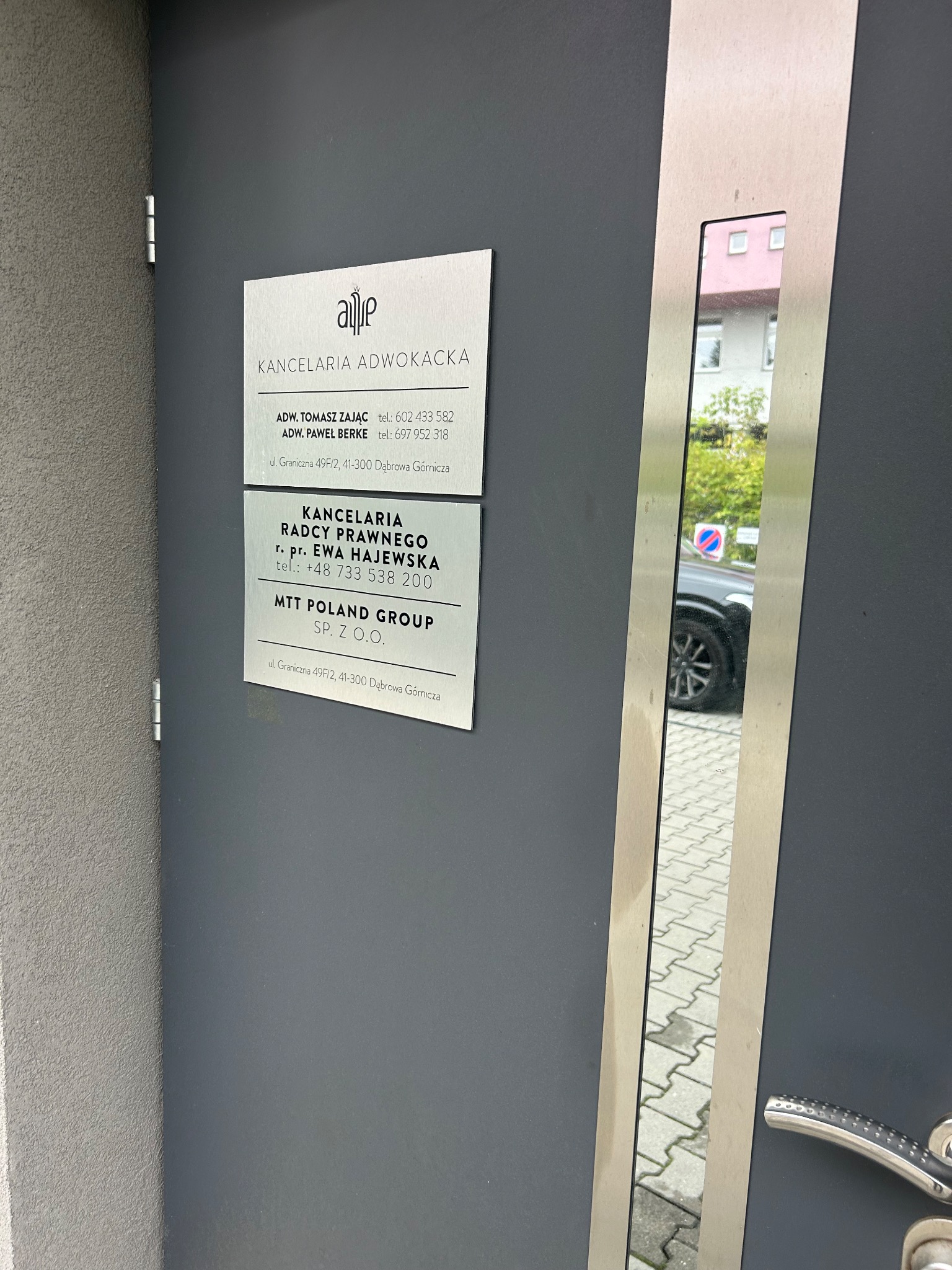 Tabliczka informacyjna na drzwiach wejściowych do kancelarii adwokackiej i radcy prawnego z nazwiskami prawników i numerami telefonów, widoczna na szarej ścianie obok metalowej listwy z odbiciem...