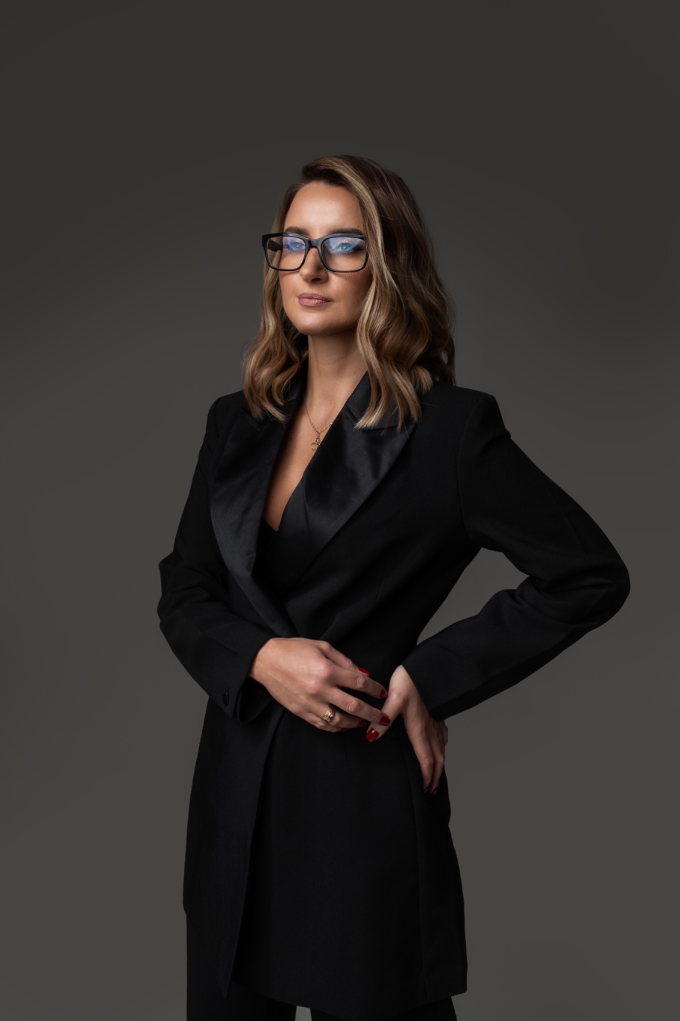 Portret kobiety w czarnym garniturze i okularach, z blond lokami, na szarym tle.