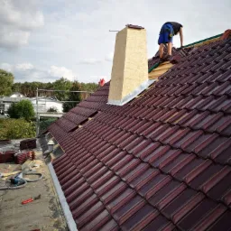 Dekarz układa bordową dachówkę ceramiczną na dachu domu jednorodzinnego, widoczny komin obłożony płytami OSB, narzędzia i zapas dachówek na dachu.