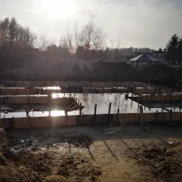 Zalane betonem szalunki pod fundamenty domu jednorodzinnego, widoczne wystające pręty zbrojeniowe, w tle ziemia i zabudowa jednorodzinna w jesiennej scenerii.