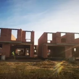 Wznoszenie ścian z czerwonej cegły na budowie dwóch domów jednorodzinnych w stanie surowym otwartym, widoczny pracownik w odblaskowej kamizelce, poranne słońce.