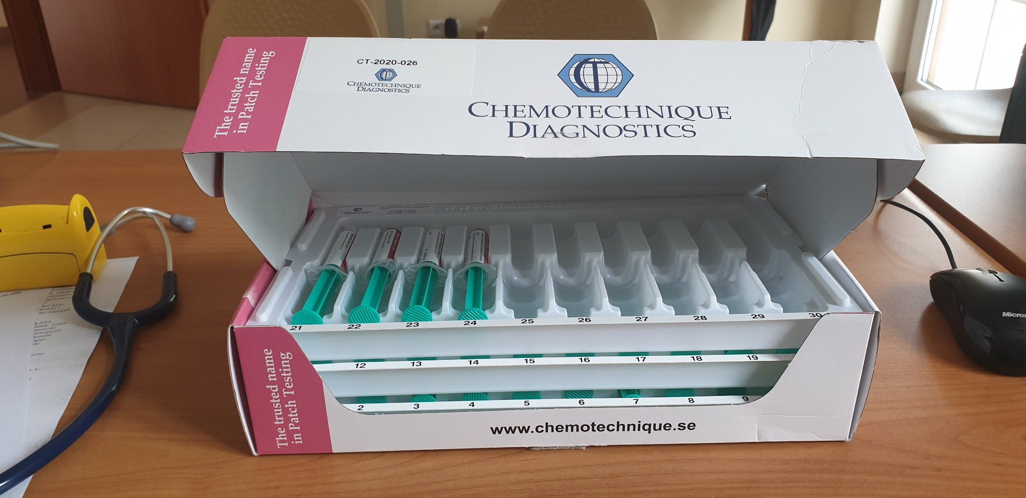 Otwarty zestaw testów płatkowych Chemotechnique Diagnostics z numerami od 1 do 30, zawierający strzykawki z zielonym płynem, leżący na drewnianym biurku obok stetoskopu i myszki komputerowej.