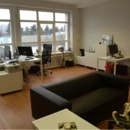 Przestronne biuro z kilkoma stanowiskami komputerowymi, widok z perspektywy niskiego stolika kawowego z dekoracyjną rośliną, duże okna z widokiem na zewnątrz.