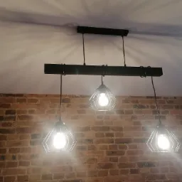 Industrialna lampa wisząca z trzema kloszami w kształcie geometrycznych klatek, zamontowana na tle ceglanej ściany, rzucająca cienie na sufit.