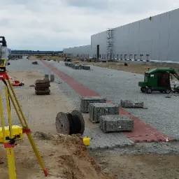 Prace drogowe w toku: widok na plac budowy z ułożoną częścią kostki brukowej, geodezyjny niwelator laserowy na statywie, maszyny budowlane i palety kostki w tle, hala magazynowa w oddali.