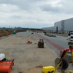 Układanie kostki brukowej na dużej powierzchni placu, widoczny sprzęt budowlany: zamiatarka, mini-ładowarka, niwelator optyczny, kostka brukowa w paletach, w tle hala magazynowa.