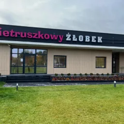 Nowoczesny budynek żłobka z dużym napisem 'Pietruszkowy Żłobek' na elewacji, otoczony zadbanym trawnikiem i nowymi nasadzeniami roślin.