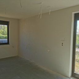 Firma Handlowo Usługowa Maciej Kaliszewski - Surowe wnętrze pokoju przed wykończeniem: ściany po wstępnym szpachlowaniu, widoczne kable zwisające z sufitu, surowa wylewka podłogowa, okno z widokiem na zieleń.