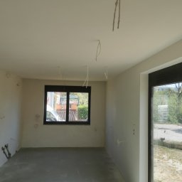 Firma Handlowo Usługowa Maciej Kaliszewski - Surowe wnętrze pokoju w trakcie remontu: ściany po szpachlowaniu, widoczne kable wystające z sufitu i ścian, okno z widokiem na zewnątrz, betonowa podłoga.