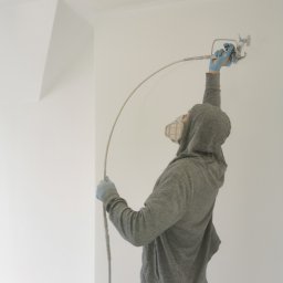 Firma Handlowo Usługowa Maciej Kaliszewski - Pracownik w masce i bluzie z kapturem maluje natryskowo sufit w pomieszczeniu w Swarożynie, używając pistoletu natryskowego.