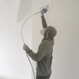 Firma Handlowo Usługowa Maciej Kaliszewski - Pracownik w masce i rękawicach natryskowo aplikuje białą farbę na ścianę w pomieszczeniu z surowym betonem na podłodze.