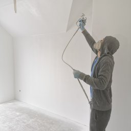 Firma Handlowo Usługowa Maciej Kaliszewski - Pracownik w masce i kapturze natryskowo aplikuje białą farbę na sufit w pustym pomieszczeniu, widoczny przewód i pistolet natryskowy.