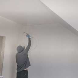Firma Handlowo Usługowa Maciej Kaliszewski - Pracownik w bluzie z kapturem, w niebieskich rękawicach, maluje natryskowo białą farbą ścianę i sufit na poddaszu, widoczny otwór drzwiowy w stanie surowym.