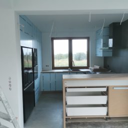 Firma Handlowo Usługowa Maciej Kaliszewski - Wnętrze kuchni w trakcie remontu; ściany przygotowane pod wykończenie, widoczne płyty izolacyjne, częściowo zamontowane szafki kuchenne z wysuwanymi szufladami, drabina malarska przy ścianie...