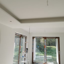 Firma Handlowo Usługowa Maciej Kaliszewski - Wnętrze w trakcie prac wykończeniowych: ściana z nałożoną masą szpachlową, sufit podwieszany z otworami na oświetlenie, drabina z narzędziami malarskimi przy oknie.