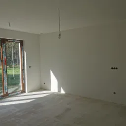 Wnętrze w stanie deweloperskim po nałożeniu gładzi, widoczne ślady po szlifowaniu na podłodze, surowa żarówka na kablu zwisająca z sufitu, okno z widokiem na zieleń.