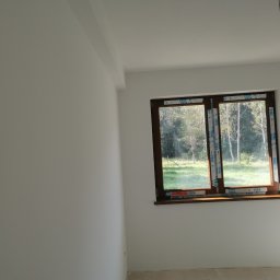 Firma Handlowo Usługowa Maciej Kaliszewski - Wnętrze pokoju po remoncie, ściany pomalowane na biało, widoczne okno z brązową ramą, zabezpieczone folią ochronną VEKA, przez okno widok na zieleń.