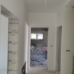 Firma Handlowo Usługowa Maciej Kaliszewski - Korytarz w trakcie prac wykończeniowych; widoczne surowe, betonowe elementy konstrukcyjne kontrastujące z świeżo pomalowanymi na biało ścianami; podłoga zabezpieczona folią malarską.
