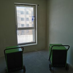 Firma Handlowo Usługowa Maciej Kaliszewski - Pokój po szpachlowaniu, zabezpieczone okno folią i taśmą, dwa zielone osuszacze kondensacyjne stoją na podłodze.