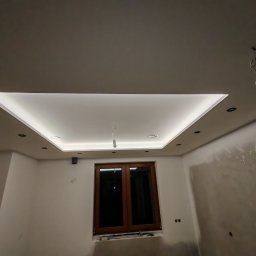 Firma Handlowo Usługowa Maciej Kaliszewski - Nowoczesny sufit podwieszany z oświetleniem LED w pomieszczeniu w trakcie prac wykończeniowych, widoczne ślady po pracach na ścianach i wisząca żarówka.