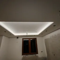 Nowoczesny sufit podwieszany z oświetleniem LED w pomieszczeniu w trakcie prac wykończeniowych, widoczne ślady po pracach na ścianach i wisząca żarówka.