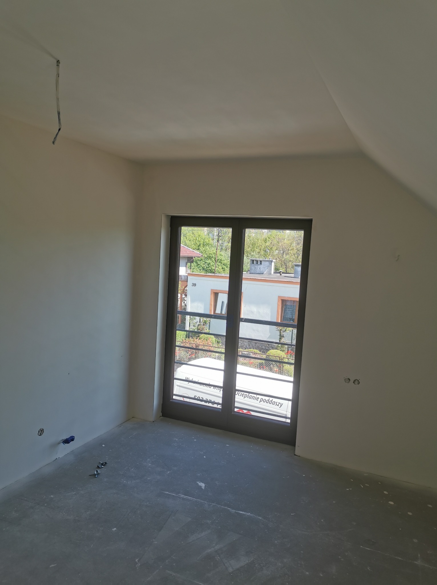 Pokój w trakcie wykańczania, ściany pomalowane na biało, surowa podłoga, widoczne wystające kable i rurki, okno z widokiem na budynek.