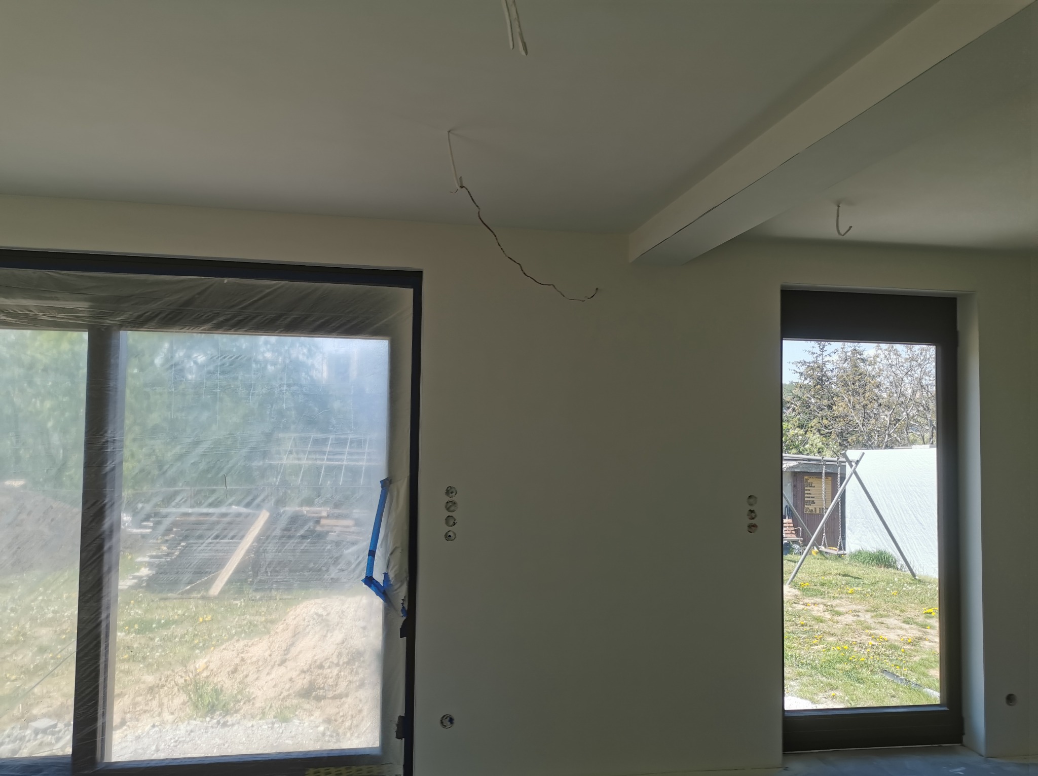 Wnętrze w trakcie prac przygotowawczych przed malowaniem: zabezpieczone okno folią, świeżo wygładzone ściany z widocznymi puszkami elektrycznymi, surowy strop z belkami.