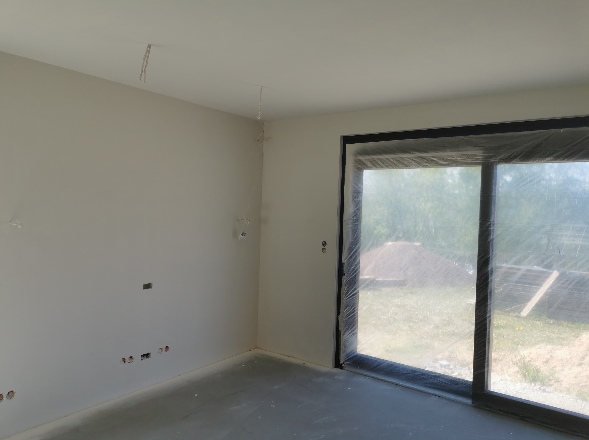Pokój w stanie deweloperskim z wygładzonymi ścianami, przygotowany do malowania, z widocznymi wyprowadzonymi przewodami elektrycznymi i zabezpieczonym folią oknem.