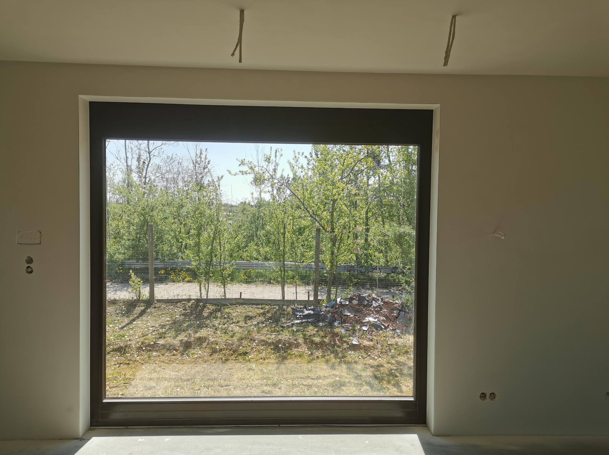 Surowe wnętrze z widokiem na zewnątrz przez duże okno; gładkie, jasne ściany i sufit w trakcie wykończenia, widoczne kable zwisające z sufitu, oraz puszki elektryczne na ścianie, przez okno widok...