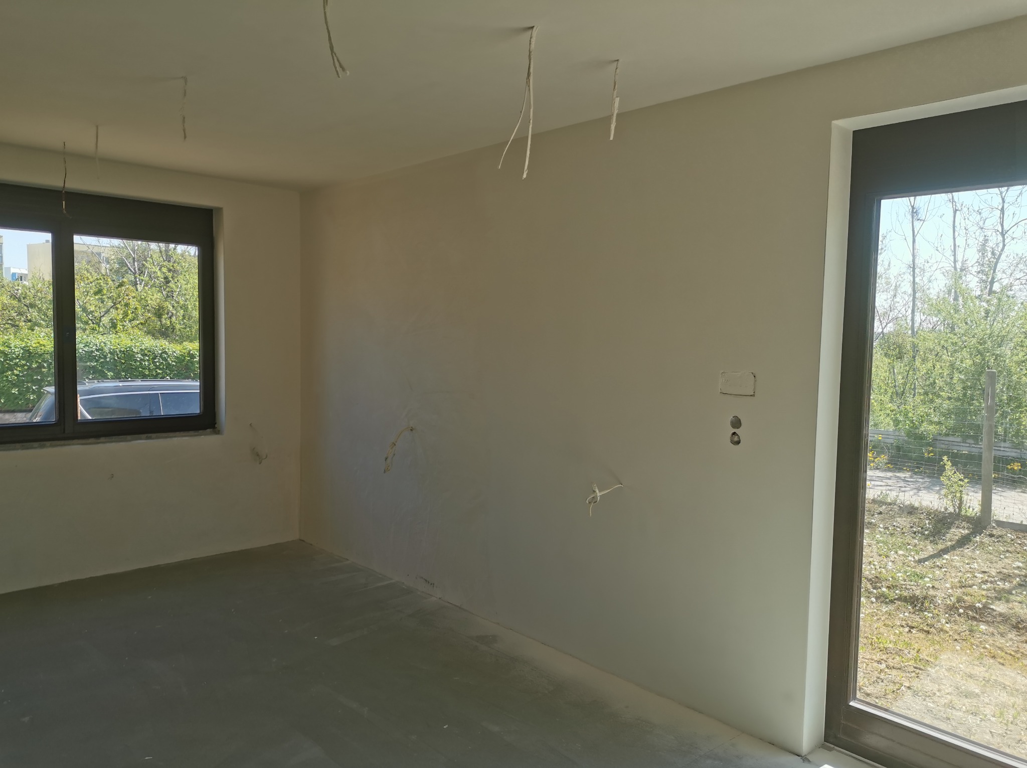 Surowe wnętrze pokoju przed wykończeniem: ściany po wstępnym szpachlowaniu, widoczne kable zwisające z sufitu, surowa wylewka podłogowa, okno z widokiem na zieleń.