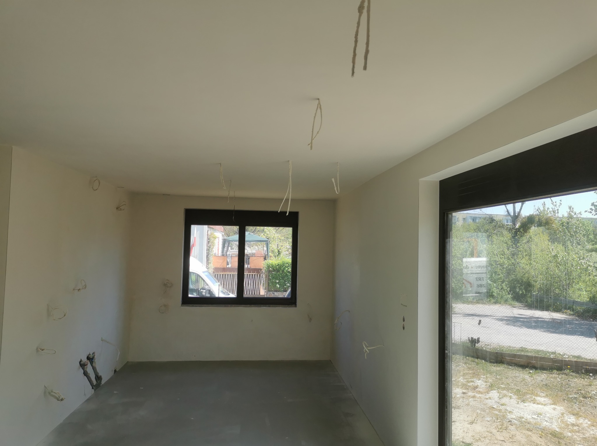 Surowe wnętrze pokoju w trakcie remontu: ściany po szpachlowaniu, widoczne kable wystające z sufitu i ścian, okno z widokiem na zewnątrz, betonowa podłoga.