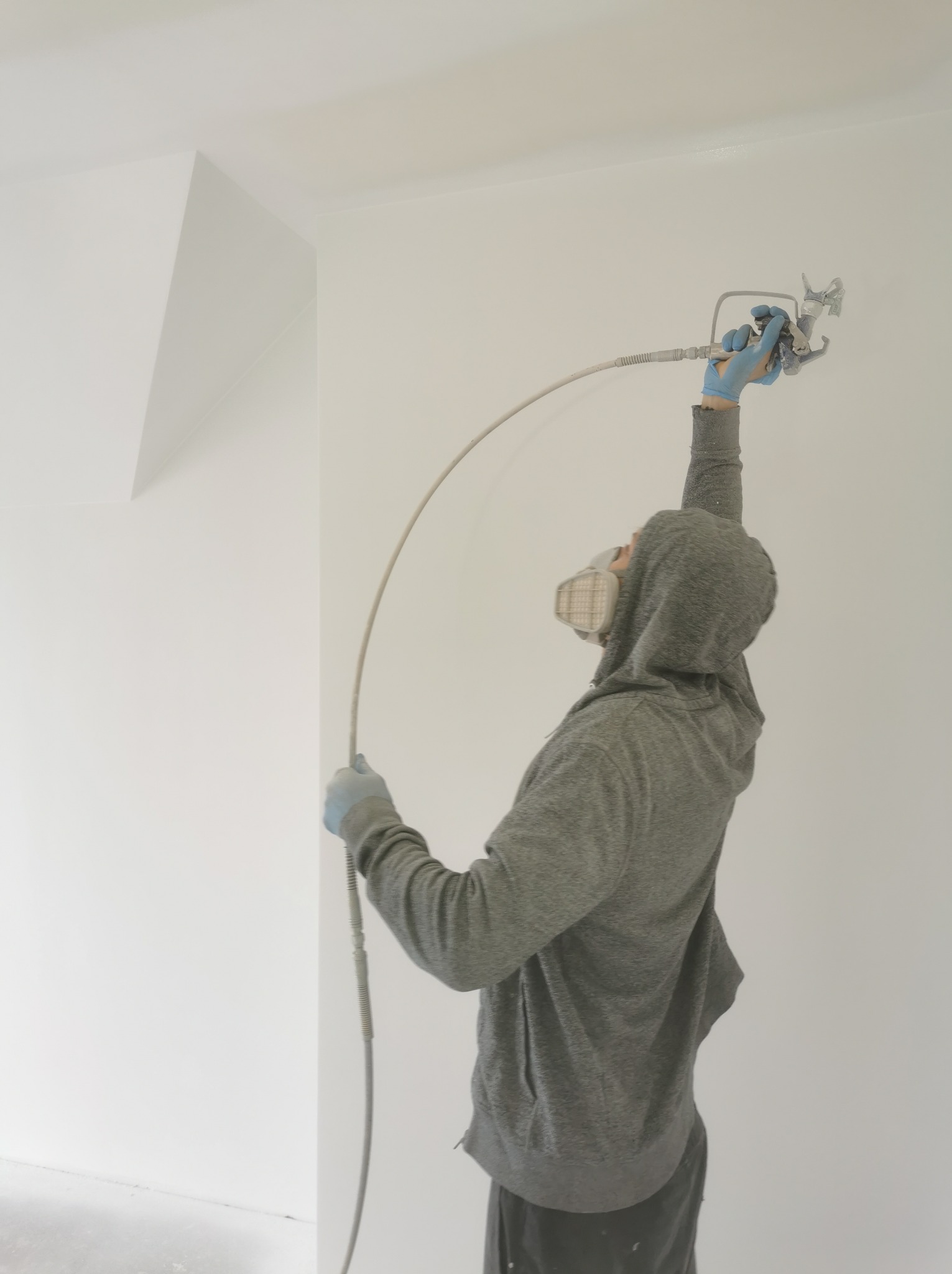 Pracownik w masce i bluzie z kapturem maluje natryskowo sufit w pomieszczeniu w Swarożynie, używając pistoletu natryskowego.