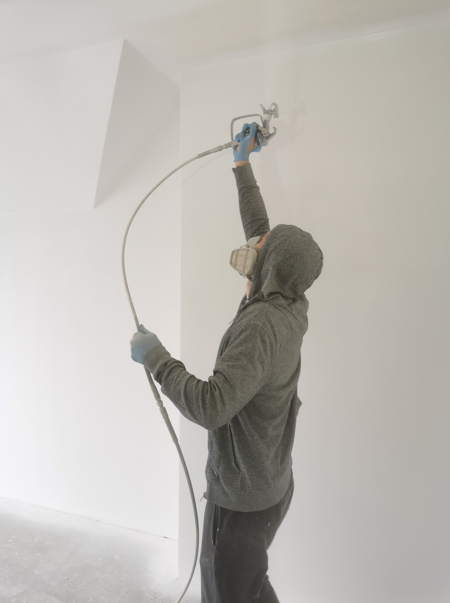 Pracownik w masce i rękawicach natryskowo aplikuje białą farbę na ścianę w pomieszczeniu z surowym betonem na podłodze.