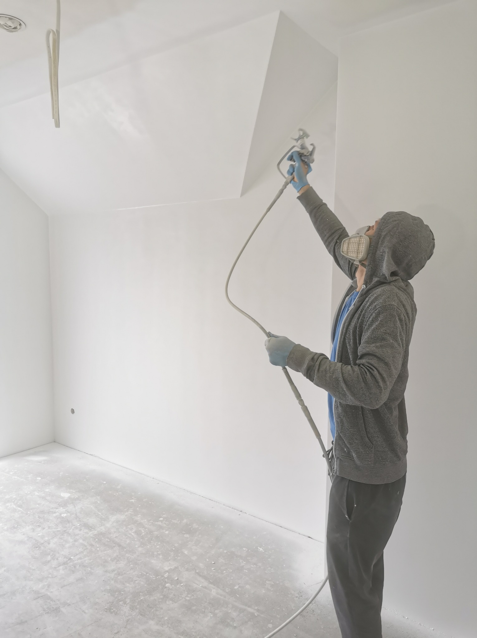 Pracownik w masce i kapturze natryskowo aplikuje białą farbę na sufit w pustym pomieszczeniu, widoczny przewód i pistolet natryskowy.