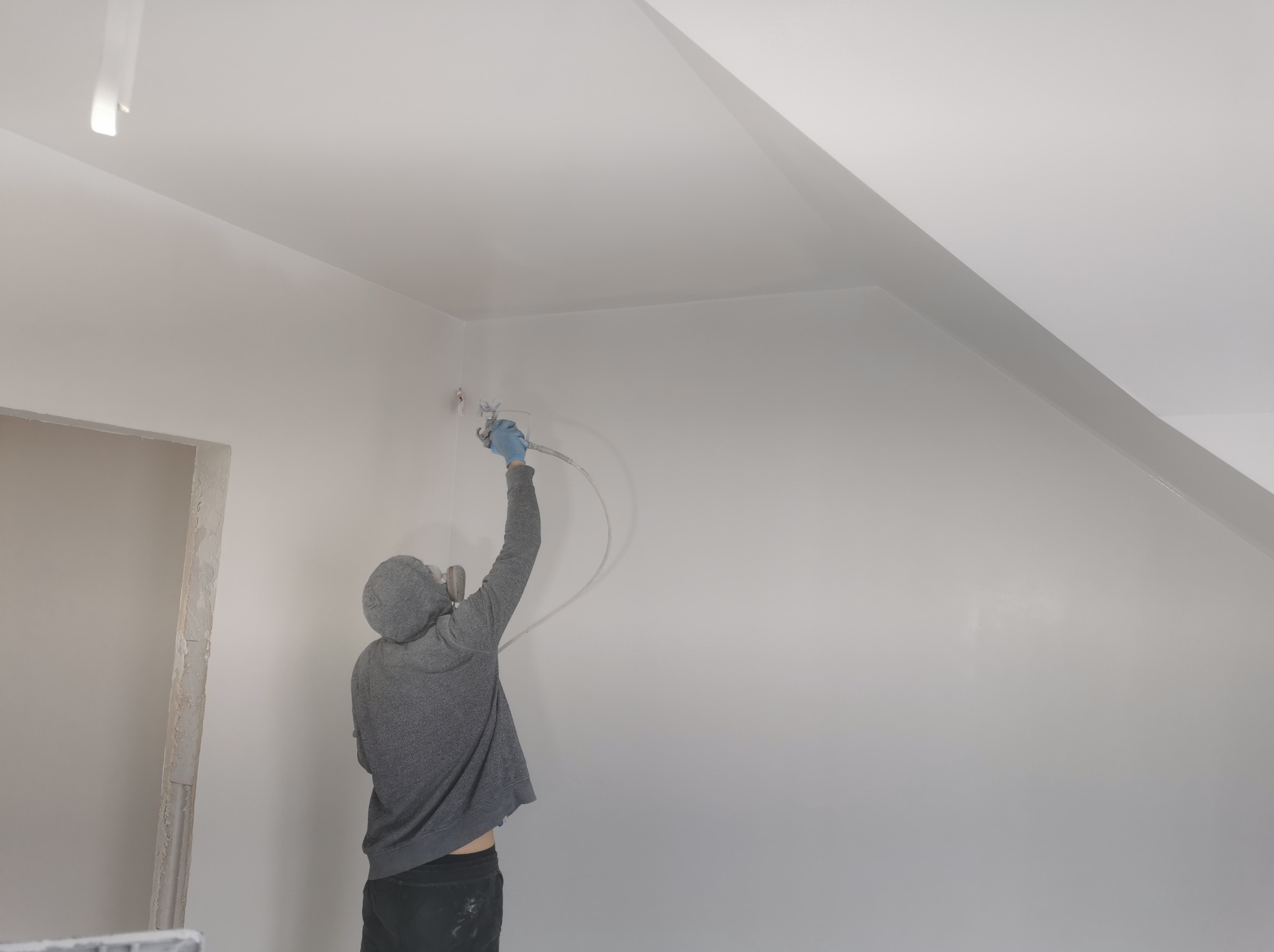 Pracownik w bluzie z kapturem, w niebieskich rękawicach, maluje natryskowo białą farbą ścianę i sufit na poddaszu, widoczny otwór drzwiowy w stanie surowym.