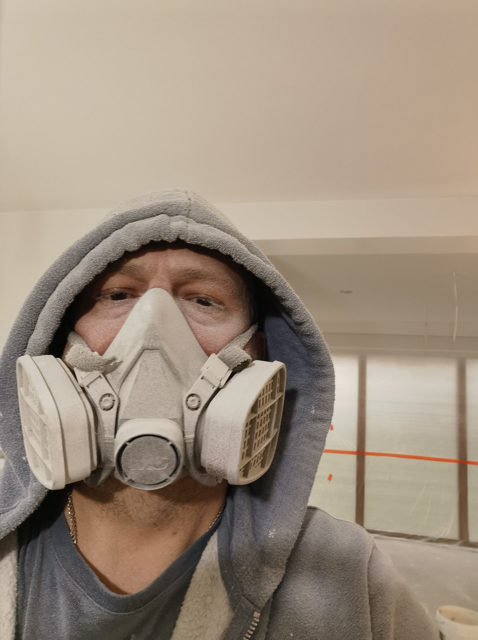 Mężczyzna w szarej bluzie z kapturem i masce ochronnej przeciwpyłowej, pokryty pyłem, w trakcie prac wykończeniowych wnętrza z widoczną taśmą malarską na ścianach.