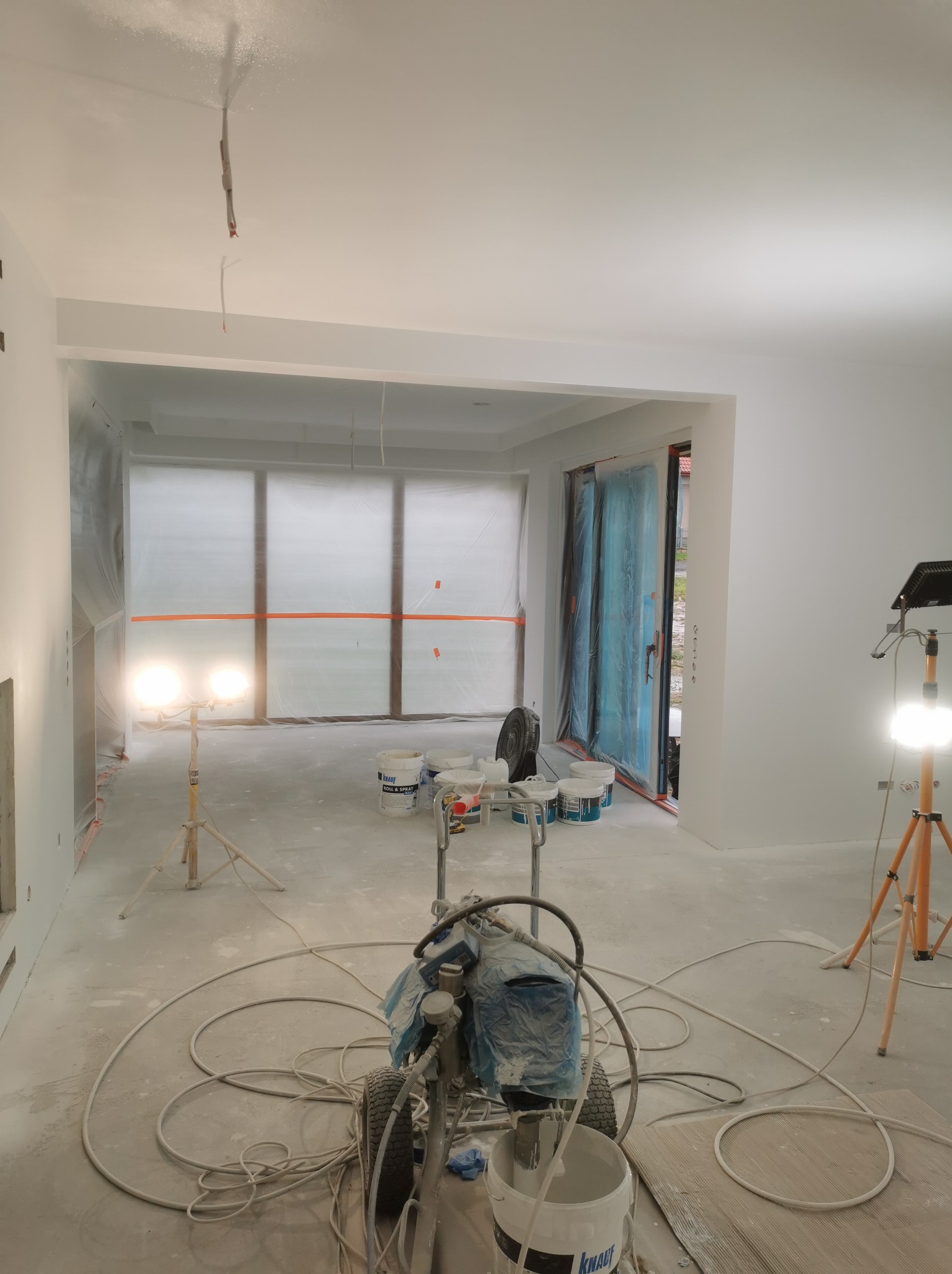 Wnętrze w trakcie prac wykończeniowych: agregat malarski na pierwszym planie, ściany i sufit świeżo pomalowane na biało, okna zabezpieczone folią, widoczne lampy budowlane i wiaderka z farbą Knauf.