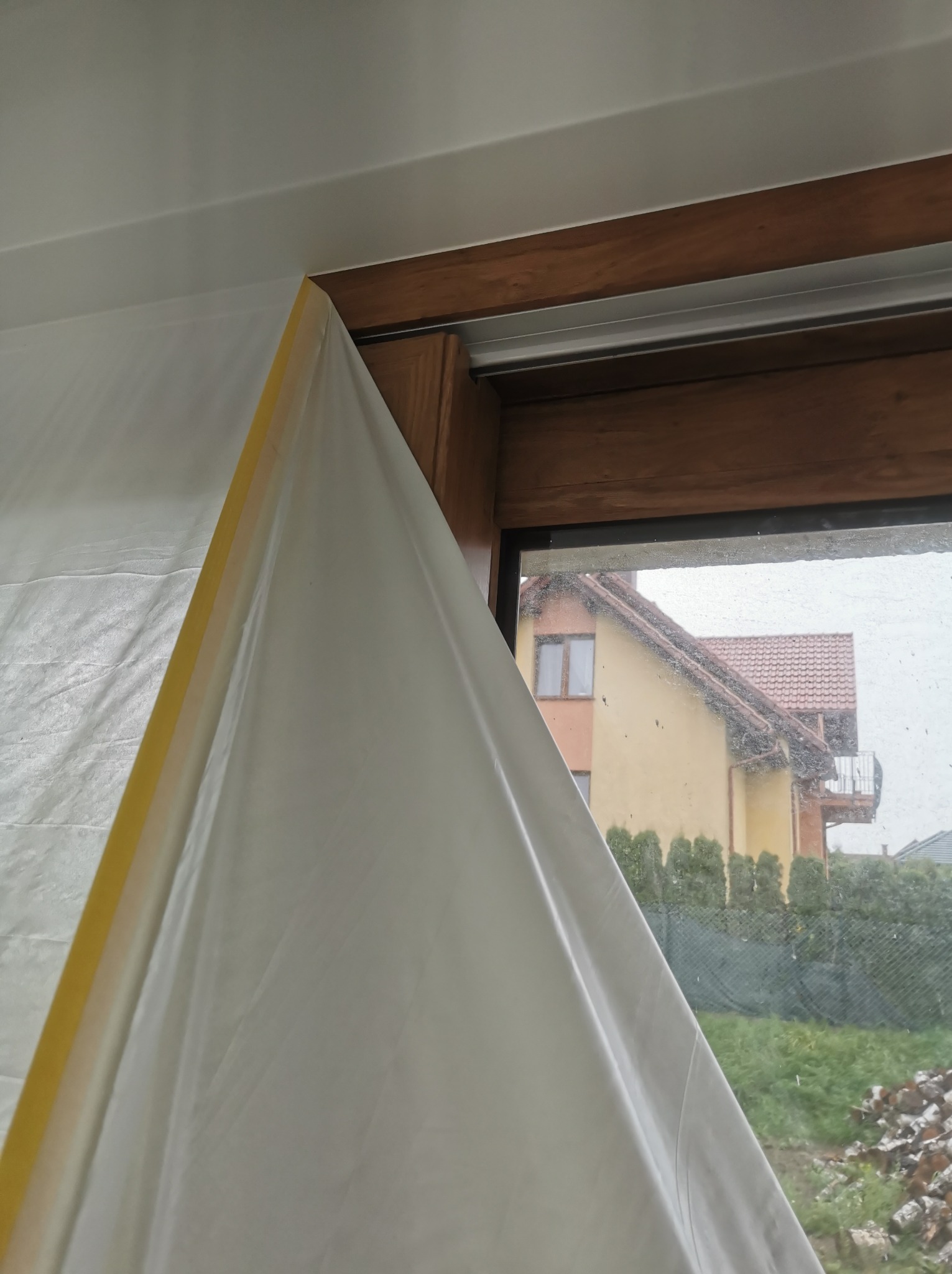 Okno zabezpieczone folią malarską przyklejoną żółtą taśmą, widok na dom z brudną szybą.