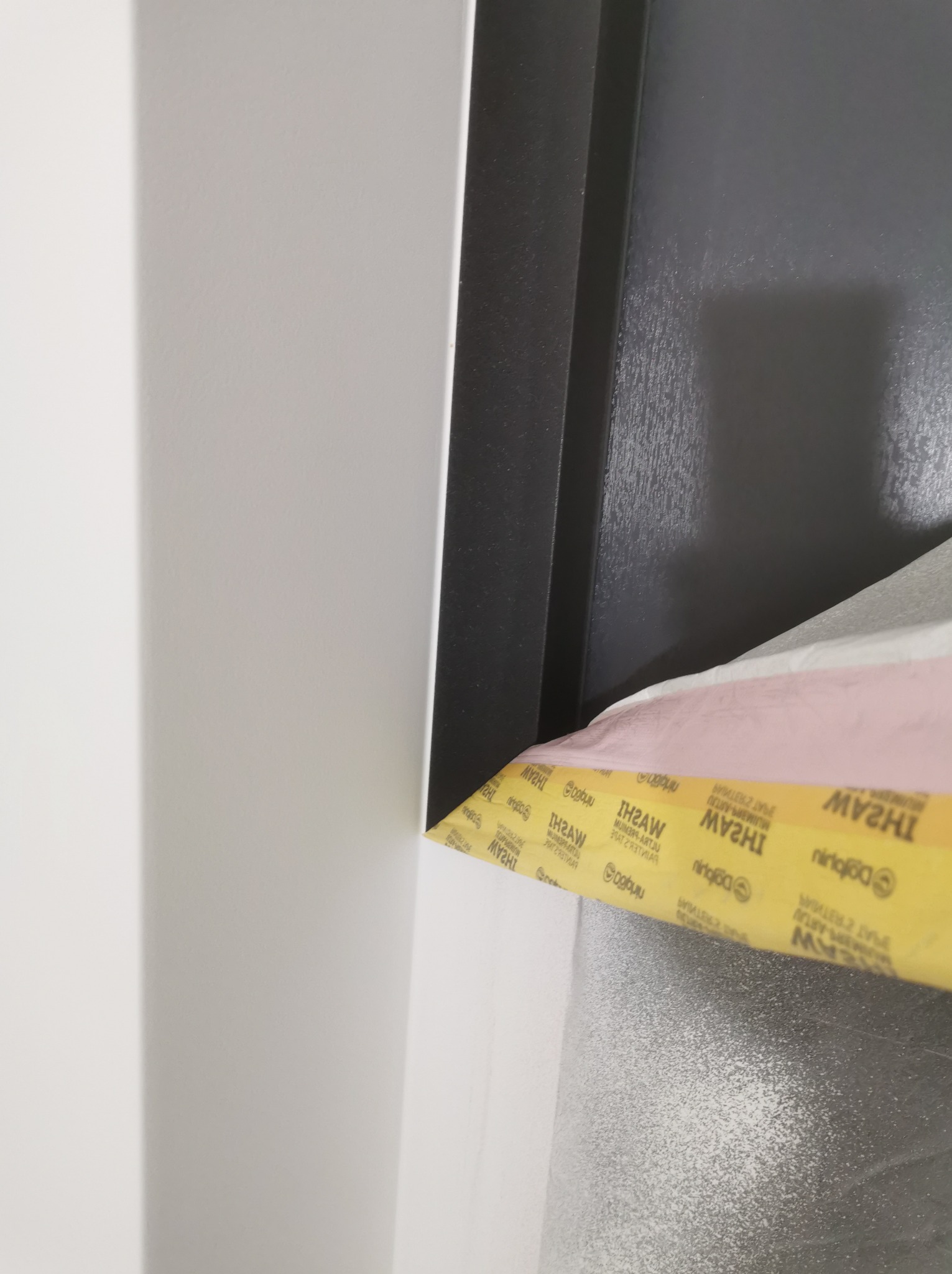 Detale przygotowania powierzchni do malowania: narożnik ściany zabezpieczony taśmą malarską z widoczną fakturą świeżo nałożonego materiału wykończeniowego.