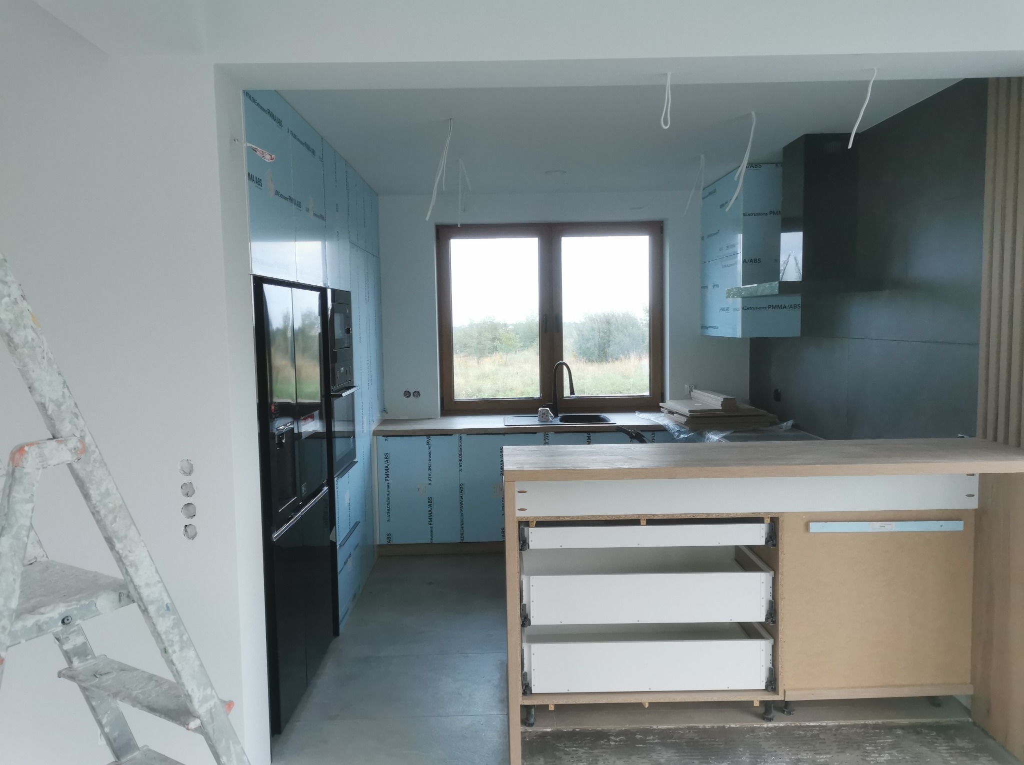 Wnętrze kuchni w trakcie remontu; ściany przygotowane pod wykończenie, widoczne płyty izolacyjne, częściowo zamontowane szafki kuchenne z wysuwanymi szufladami, drabina malarska przy ścianie...