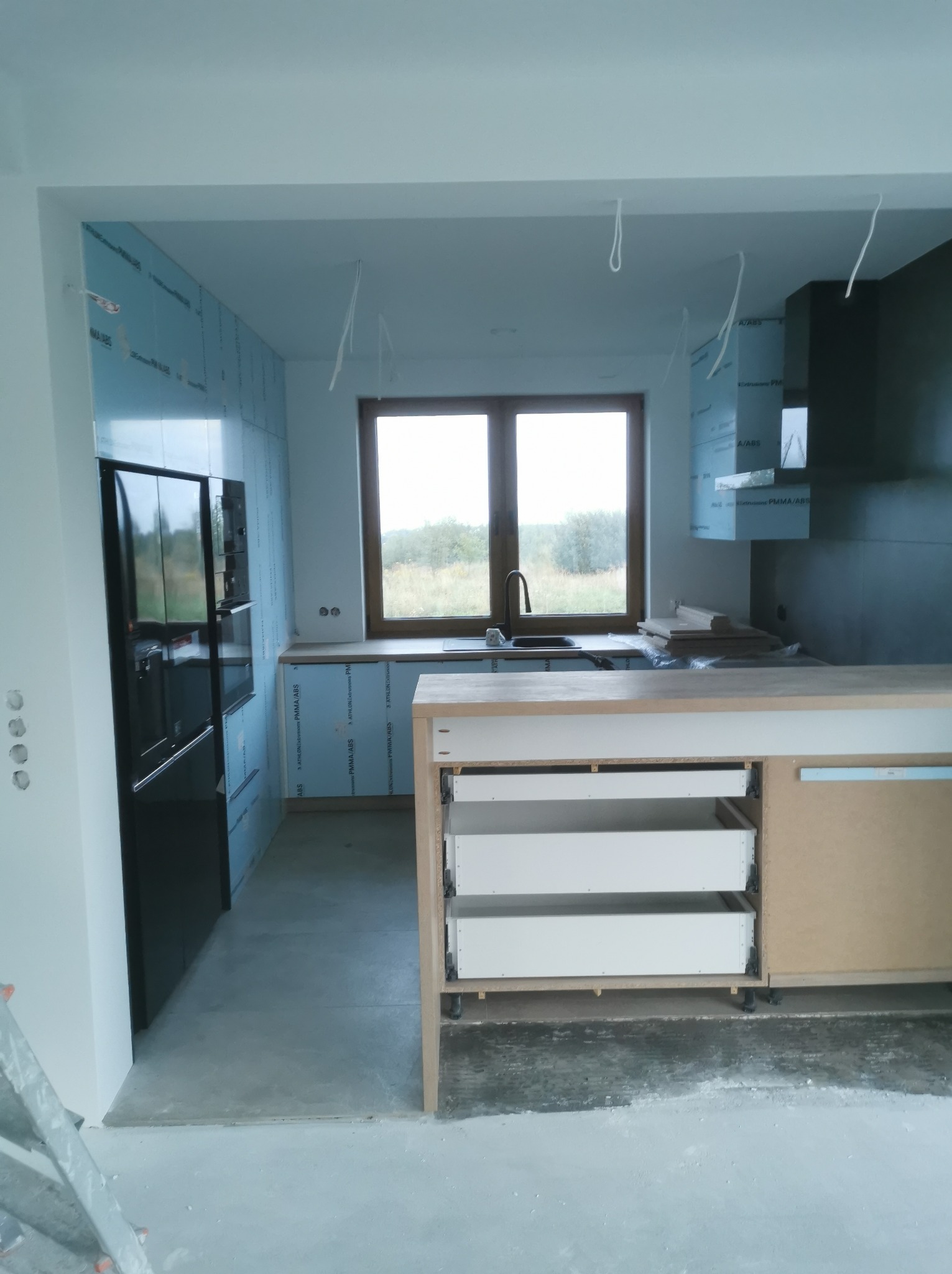 Wnętrze kuchni w trakcie remontu, ściany przygotowane pod wykończenie, widoczne niezabudowane szafki kuchenne z szufladami, fragment drabiny malarskiej.