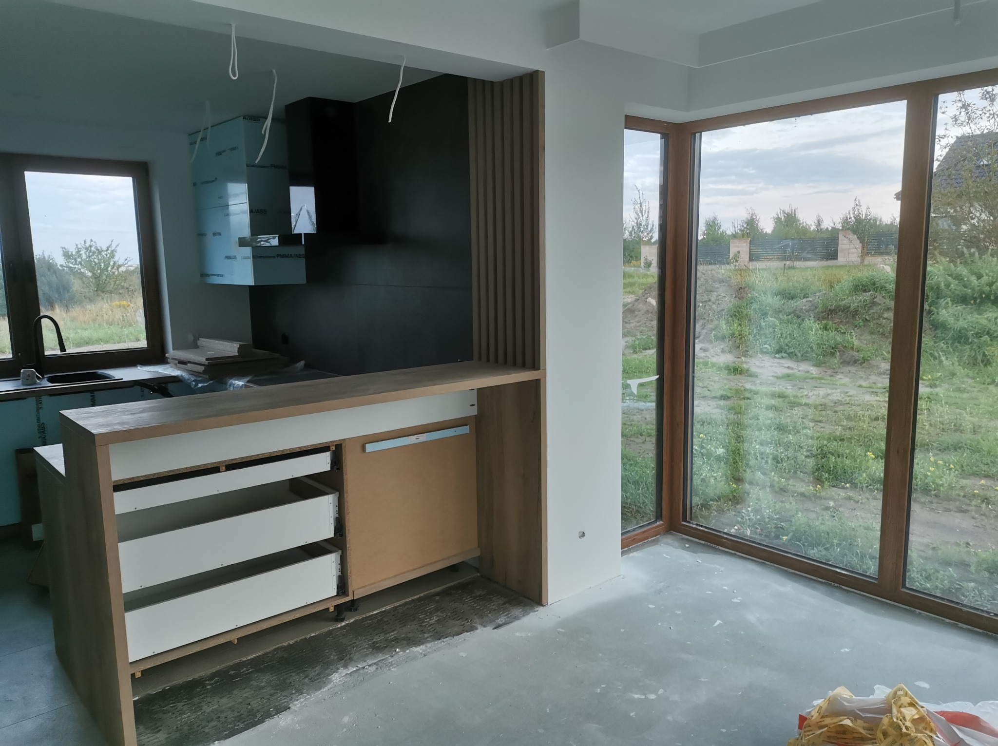 Nowoczesna kuchnia w trakcie wykończenia: widoczne niezabezpieczone elementy, surowe ściany i podłoga betonowa, okna panoramiczne z widokiem na zieleń.