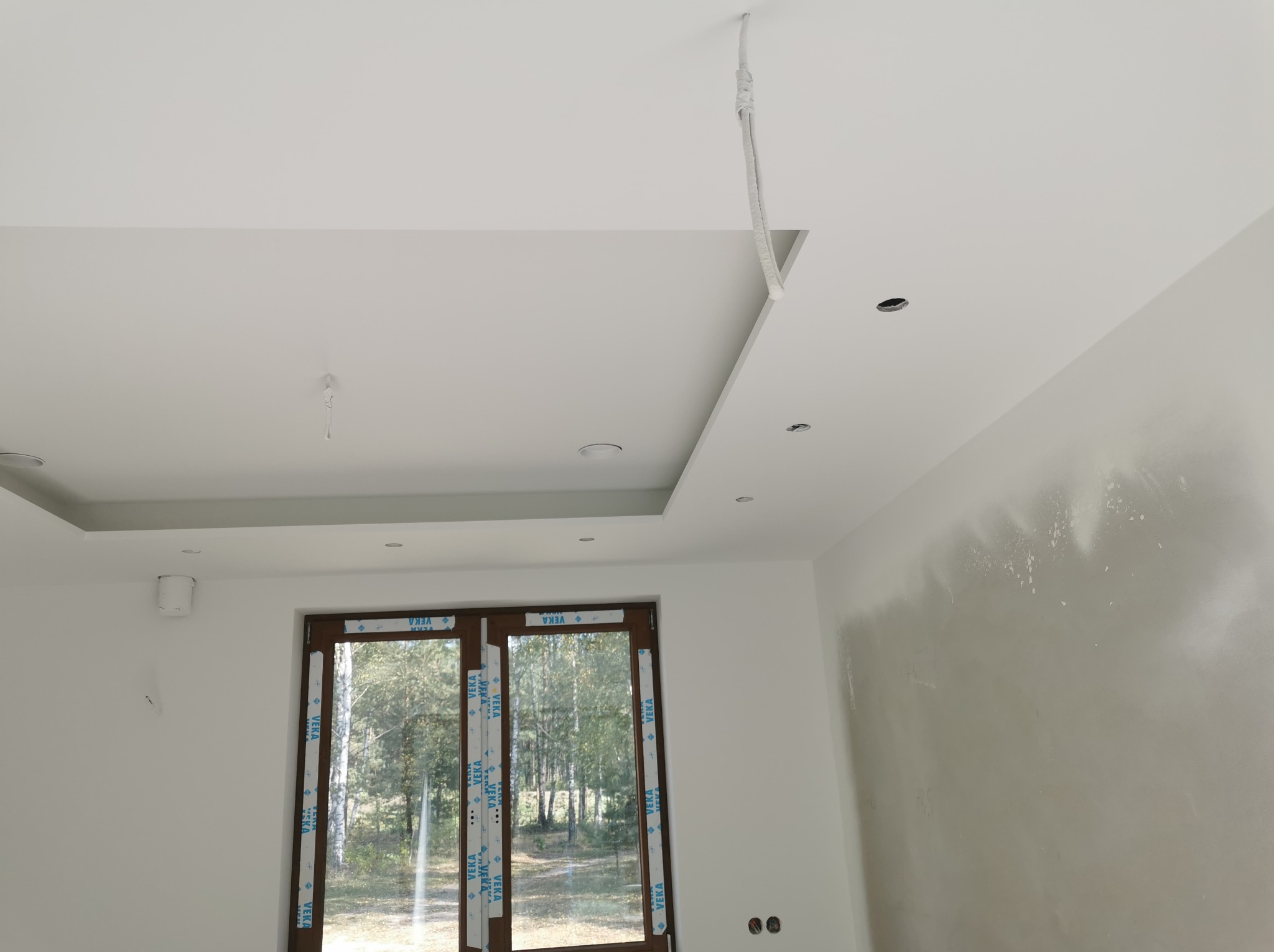 Świeżo pomalowane wnętrze z widocznymi śladami szpachlowania na ścianie, dekoracyjnym sufitem podwieszanym z oświetleniem punktowym i wystającymi kablami, okno z naklejkami producenta.