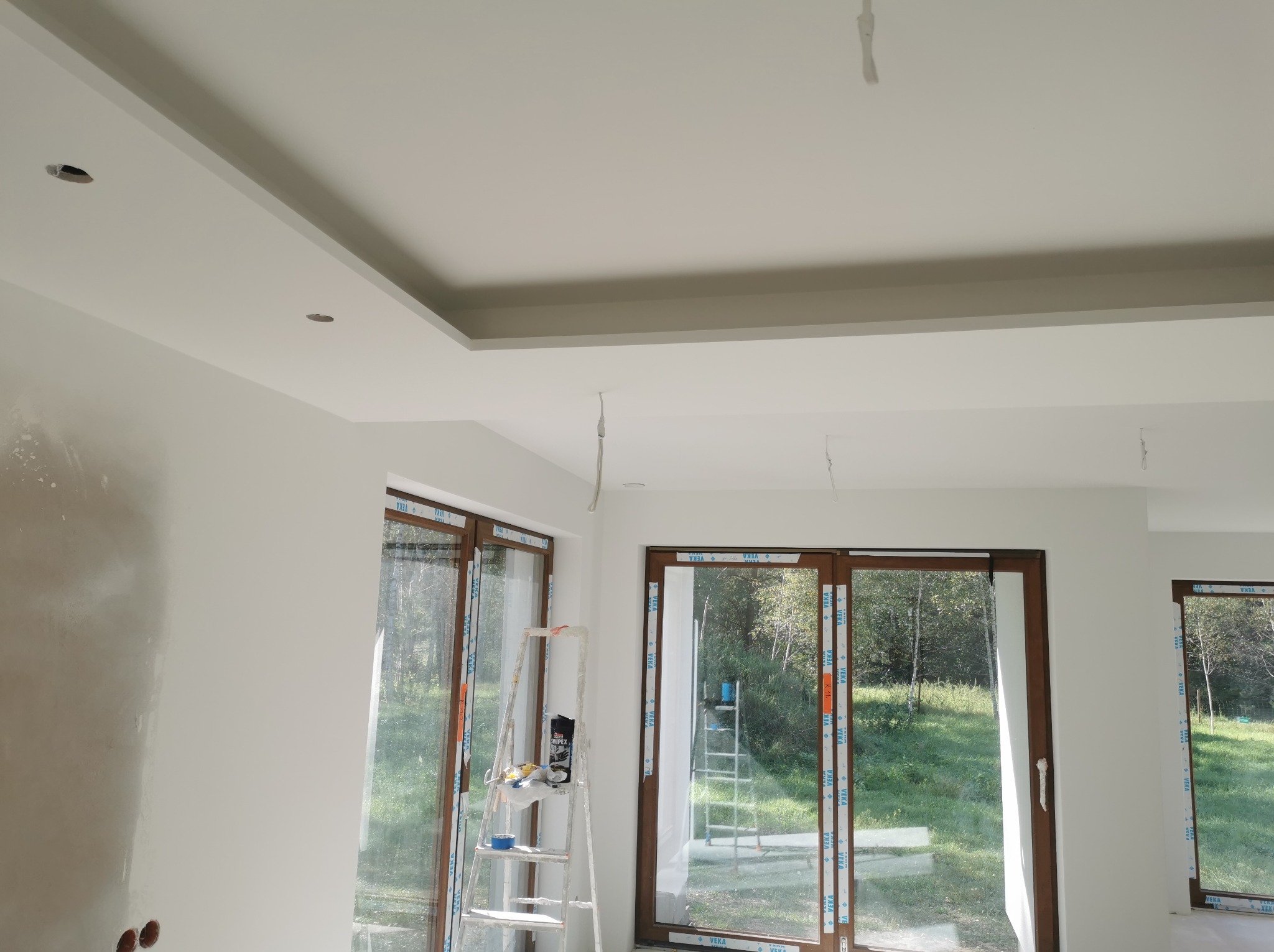 Wnętrze w trakcie prac wykończeniowych: ściana z nałożoną masą szpachlową, sufit podwieszany z otworami na oświetlenie, drabina z narzędziami malarskimi przy oknie.