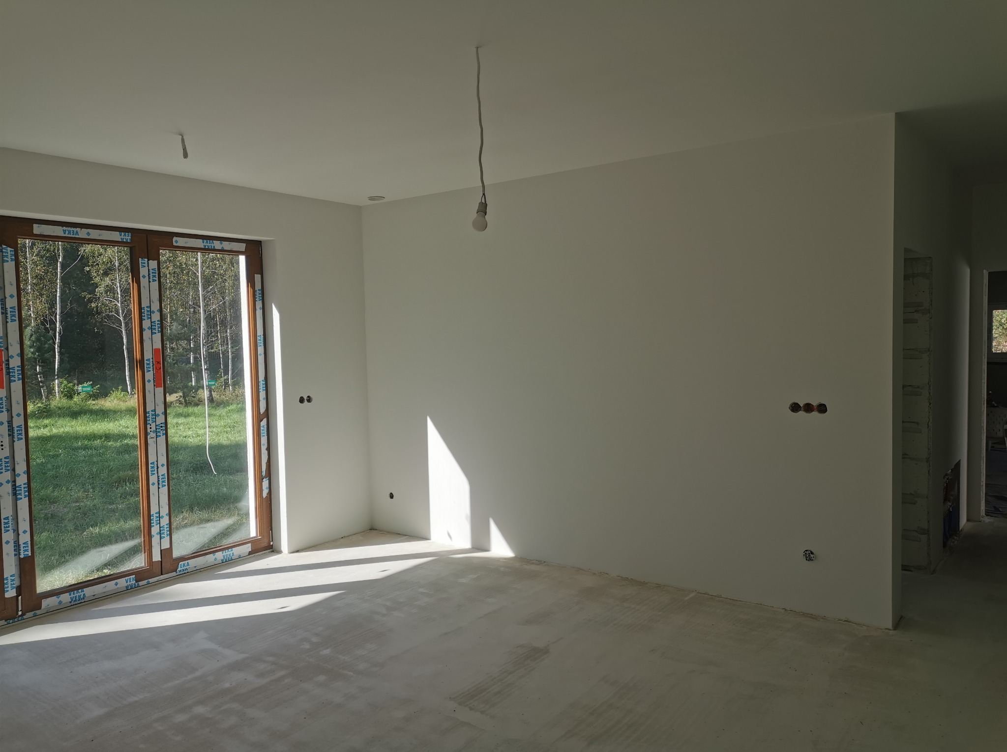 Wnętrze w stanie deweloperskim po nałożeniu gładzi, widoczne ślady po szlifowaniu na podłodze, surowa żarówka na kablu zwisająca z sufitu, okno z widokiem na zieleń.