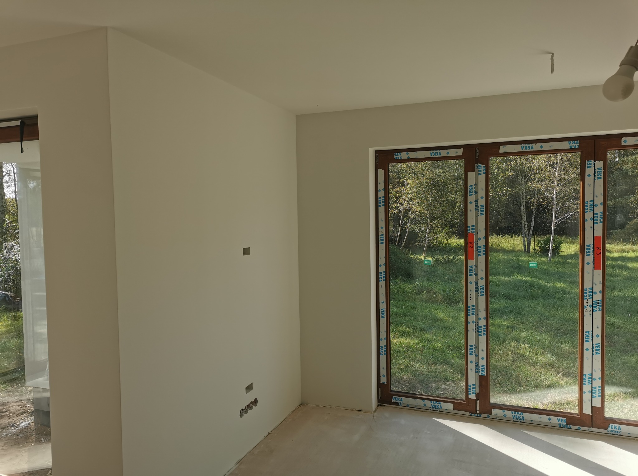 Wnętrze pokoju po remoncie, ściany wykończone na gładko, widoczne okno tarasowe z widokiem na zieleń, surowy beton na podłodze.