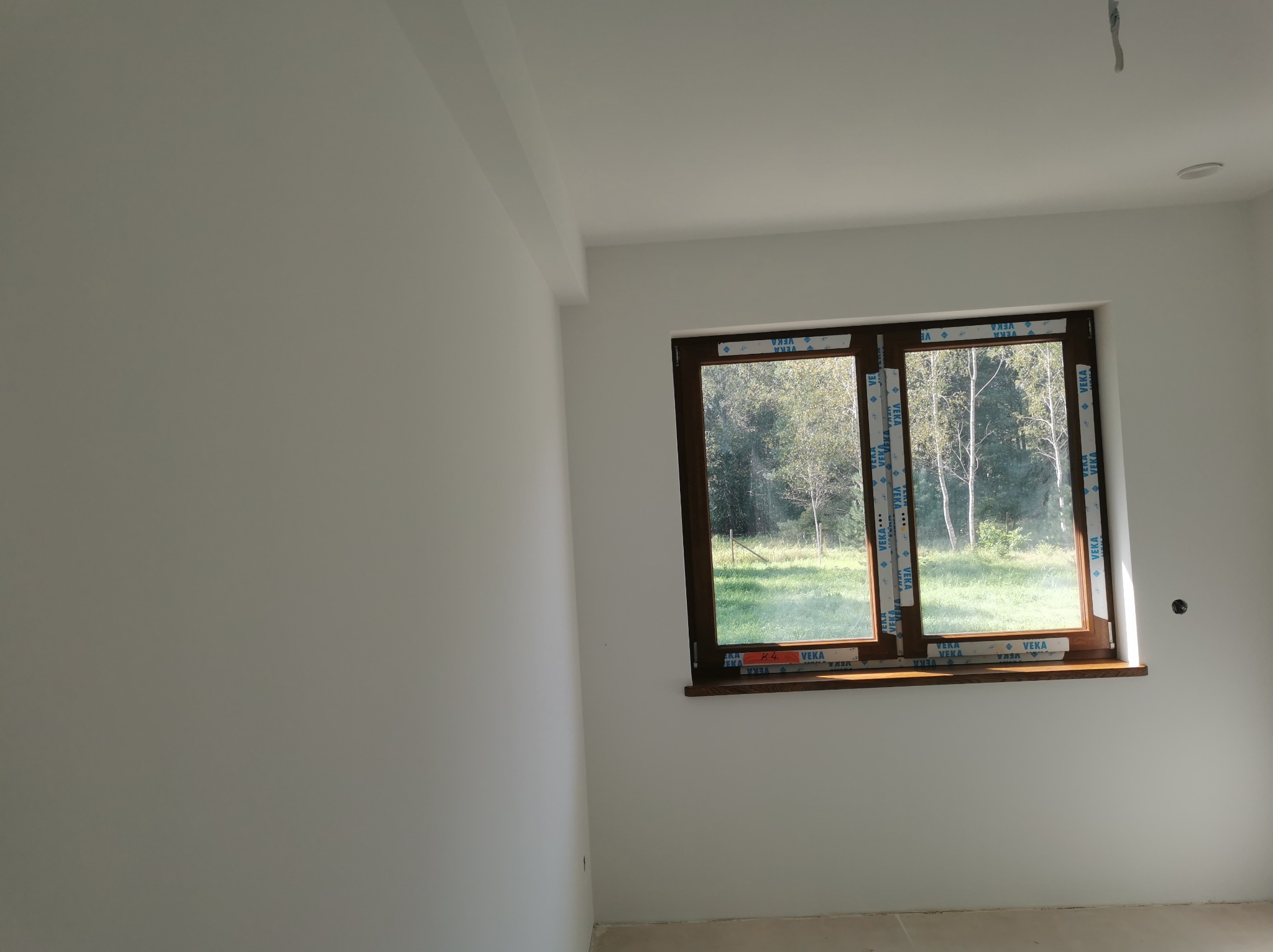 Wnętrze pokoju po remoncie, ściany pomalowane na biało, widoczne okno z brązową ramą, zabezpieczone folią ochronną VEKA, przez okno widok na zieleń.